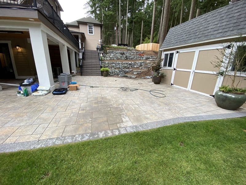 Patio renovation with stone pavers, landscaping, and a nearby shed in a lush backyard.