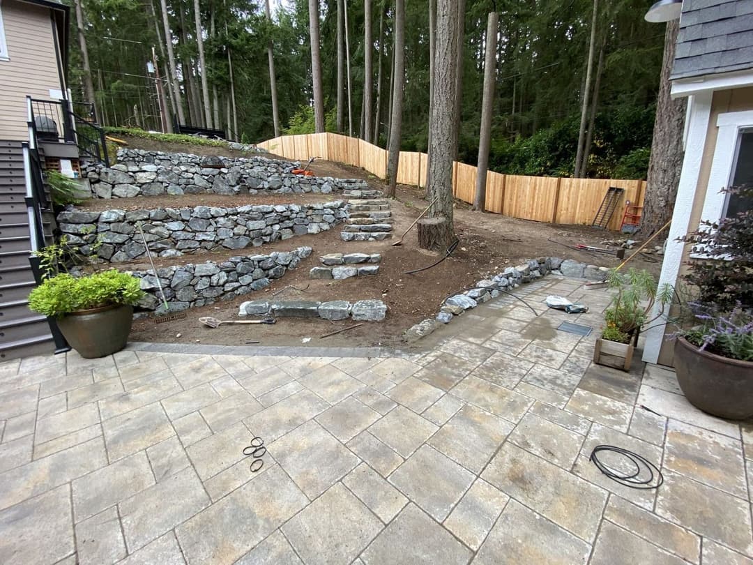 Landscaped yard with stone steps, retaining wall, and wooden fence in a forested area.