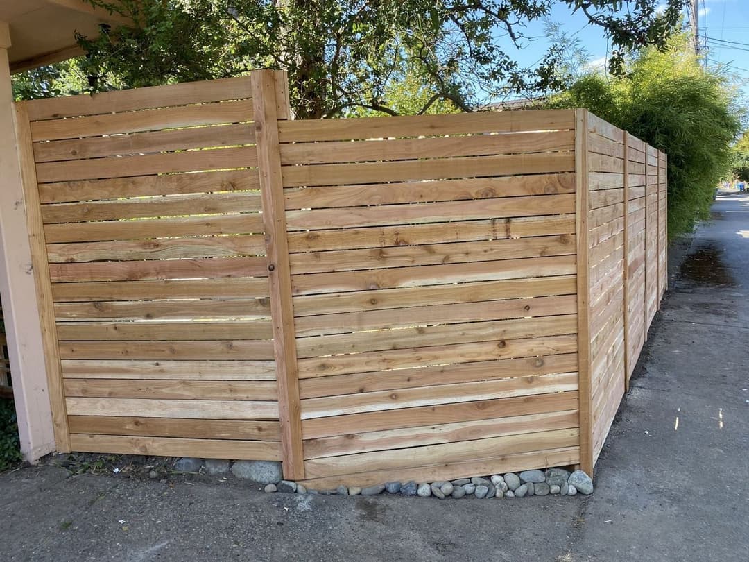 Wooden slatted fence with a natural finish in a residential area, surrounded by greenery.