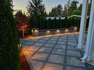 Modern outdoor patio with stone wall and ambient lighting surrounded by tall trees.