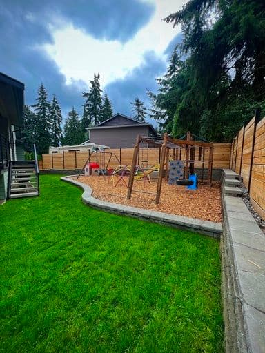 Playground with swings and climbing structure in a yard surrounded by trees and wooden fence.