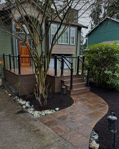 House exterior with modern deck, black railing, and stone pathway surrounded by greenery.